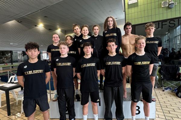 Gruppe von Schwimmern in schwarzen T-Shirts mit &bdquo;Karlsruhe&ldquo; sammeln sich auf einem Podest in einem Hallenbad.