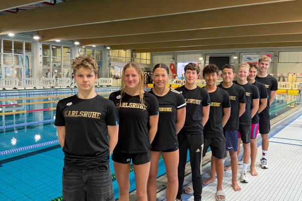 Gruppe junger Schwimmer in schwarzen T-Shirts mit &bdquo;Karlsruhe&ldquo; am Rand eines Schwimmbeckens.