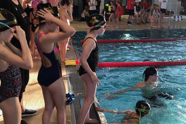 Kinder in Schwimmbekleidung stehen am Beckenrand, w&auml;hrend im Wasser schwimmende Teilnehmer trainieren.