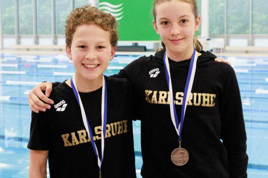 Zwei junge Schwimmer aus Karlsruhe mit Medaillen stehen in einem Schwimmbad, im Hintergrund sind Wettkampfbahnen sichtbar.