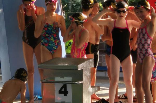 Kinder in Schwimmbekleidung stehen am Beckenrand, bereit f&uuml;r einen Wettkampf, mit einem Startblock im Vordergrund.