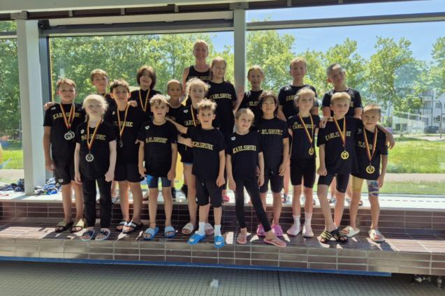 Gruppe von Kindern in Sportkleidung mit Medaillen, posierend vor einem Fenster in einem Schwimmbad.