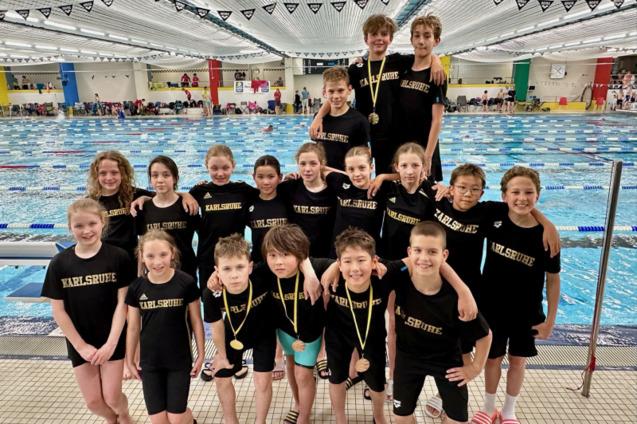 Kinder-Schwimmteam aus Karlsruhe posiert stolz mit Medaillen vor einem Schwimmbecken.