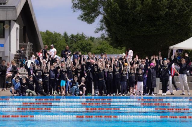 Gruppenselfie von Kindern und Erwachsenen am Schwimmbad, alle mit erhobenen H&auml;nden und Medaillen um den Hals.