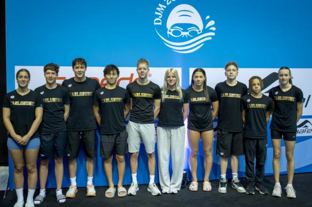 Gruppenfoto von zehn Schwimmern in schwarzen T-Shirts mit der Aufschrift &bdquo;Karlsruhe&ldquo; vor einer blauen Hintergrundwand.