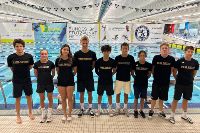 Gruppe junger Schwimmer in schwarzen T-Shirts mit &bdquo;Karlsruhe&ldquo;-Aufdruck am Beckenrand eines Schwimmbads.