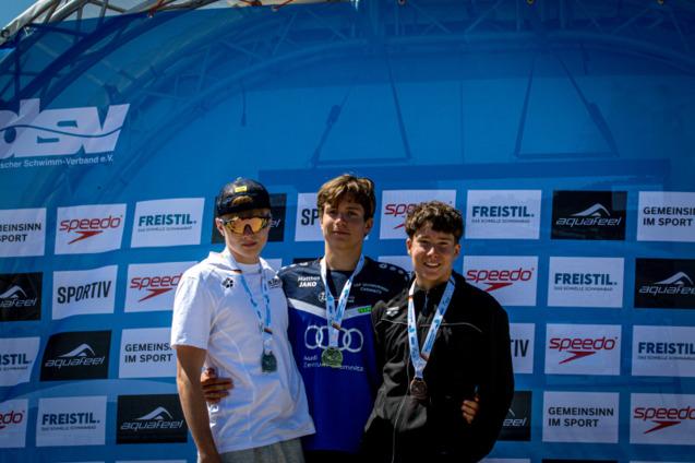 Drei junge Schwimmer stehen l&auml;chelnd auf dem Siegertreppchen, jeder mit Medaille vor einem Sponsorenbanner.