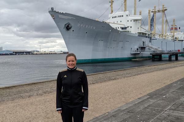 Frau in Sportkleidung steht am Ufer mit dem Frachtschiff "Dresden" im Hintergrund und bew&ouml;lktem Himmel.