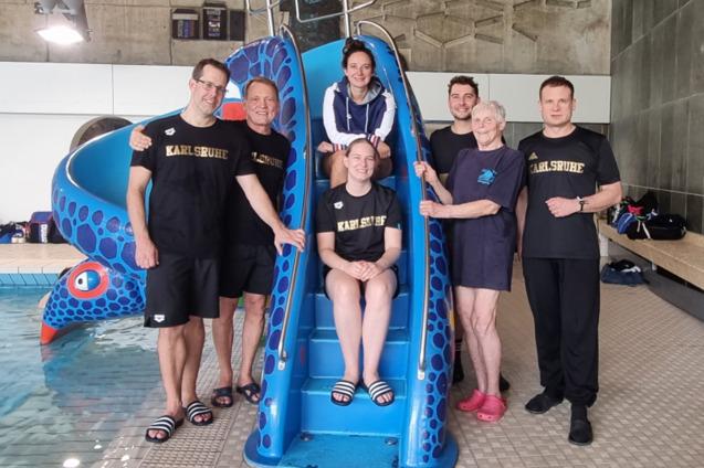 Gruppe von sechs Personen vor einer blauen Wasserrutsche in einem Hallenbad, l&auml;chelnd und in Sportkleidung.