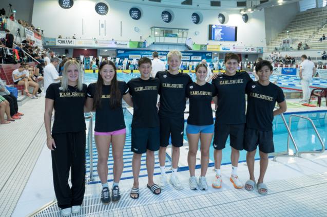 Gruppe von Schwimmern in einheitlichen schwarzen Shirts mit dem Aufdruck "Karlsruhe" vor einem Wettkampfbecken.