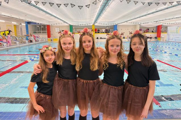 F&uuml;nf M&auml;dchen in schwarzen T-Shirts und braunen Tutus posieren am Beckenrand eines Schwimmbads mit Blumenkr&auml;nzen im Haar.