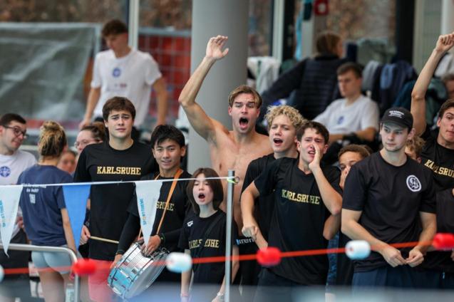 Gruppe junger Schwimmer aus Karlsruhe feuert begeistert am Beckenrand an, einige tragen Trikots der Mannschaft.
