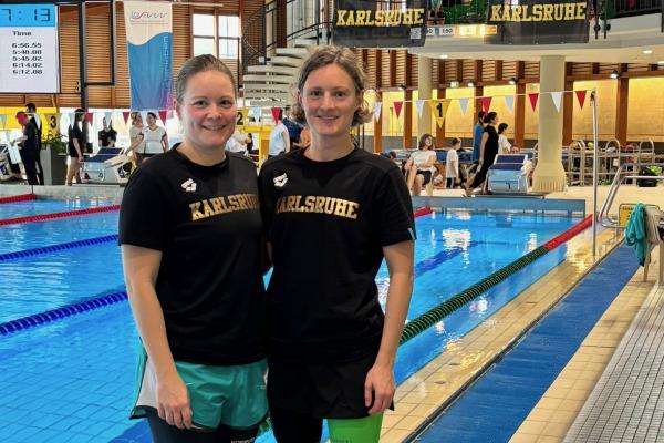 Zwei Schwimmerinnen in Sportbekleidung stehen am Beckenrand eines Schwimmbads mit Wettkampf-Atmosphäre.