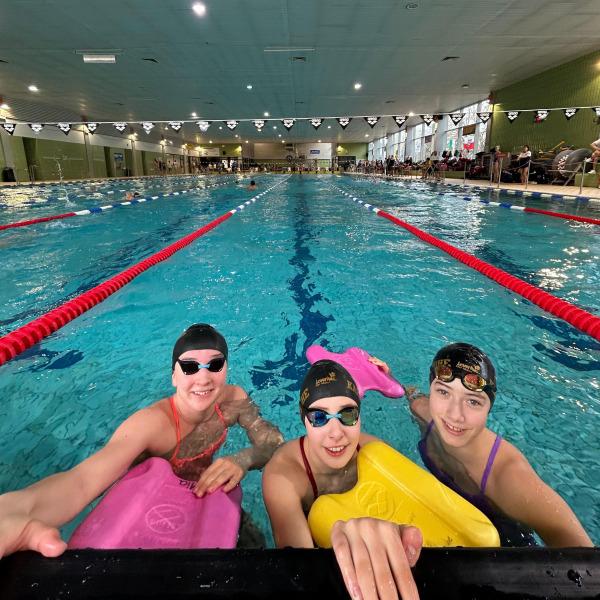 Drei schwimmende Kinder mit Schwimmbrettern posieren am Beckenrand eines Hallenbades.