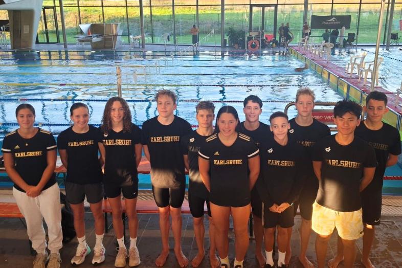 Gruppe von zw&ouml;lf Schwimmern in schwarzen T-Shirts mit &bdquo;Karlsruhe&ldquo; vor einem Wettkampfbecken.
