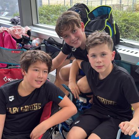 Drei Jungen in Sportkleidung sitzen lächelnd auf Campingstühlen in einer Sporteinrichtung.