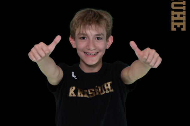 Junge mit Daumen hoch, tr&auml;gt schwarzes Shirt mit Schriftzug &bdquo;Karlsruhe&ldquo;, Hintergrund mit Text zu Schwimmmeisterschaften.