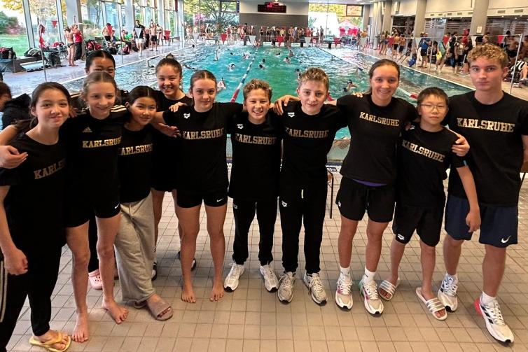 Gruppenshot von Schwimmern in einheitlichen schwarzen Shirts mit "Karlsruhe" vor einem Schwimmbecken mit Wettk&auml;mpfen.