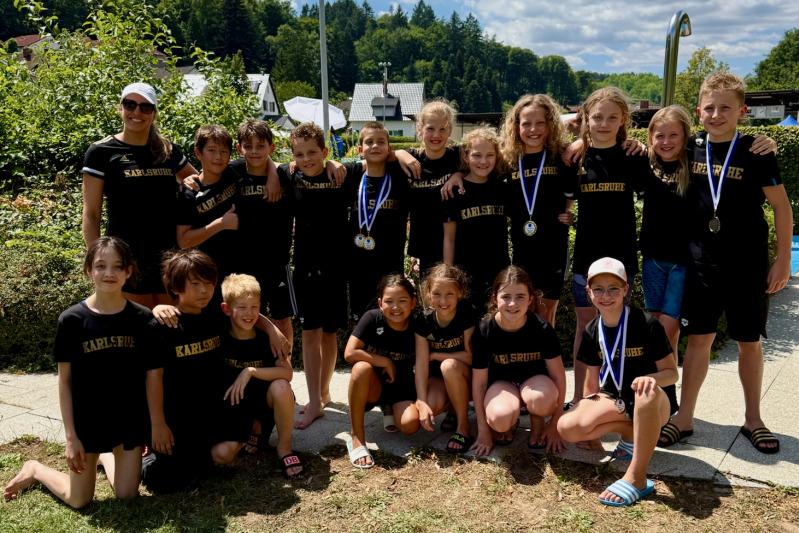 Gruppe von Kindern in schwarzen Shirts mit Aufschrift "Karlsruhe", posierend mit Medaillen unter blauem Himmel.