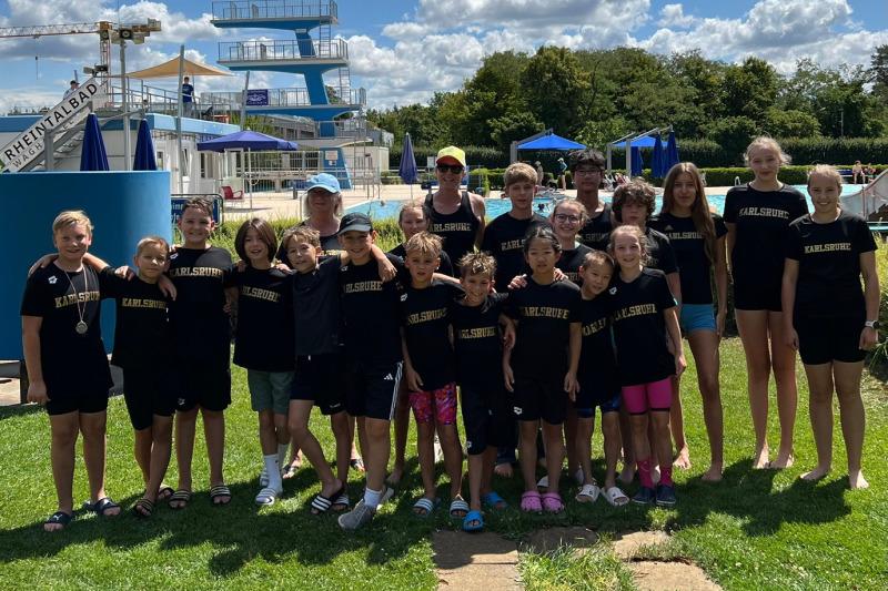 Gruppe von Kindern und Betreuern in einheitlichen T-Shirts vor einem Schwimmbad mit Sprungturm und blauen Liegen.