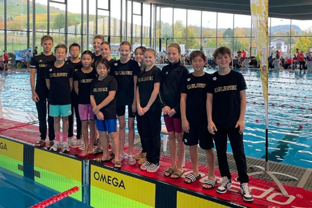 Gruppe junger Schwimmer in einheitlichen T-Shirts mit &bdquo;Karlsruhe&ldquo; stehen am Beckenrand in einem modernen Schwimmbad.