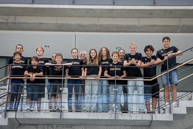 Gruppe von 13 Jugendlichen in schwarzen T-Shirts auf einer Galerie, lächelnd und an einem Geländer stehend.