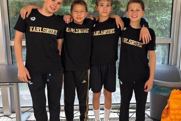 Vier Kinder stehen l&auml;chelnd zusammen in schwarzen T-Shirts mit der Aufschrift "Karlsruhe" vor einem Fenster.