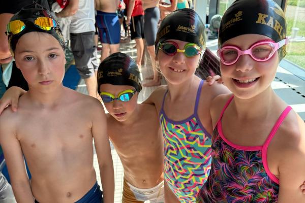 Vier Kinder in Badebekleidung und Schwimmkappen stehen zusammen am Poolrand, fr&ouml;hlich l&auml;chelnd f&uuml;r ein Foto.