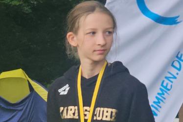 M&auml;dchen in schwarzem Hoodie mit Medaille steht vor Zelt und Banner im Freien.